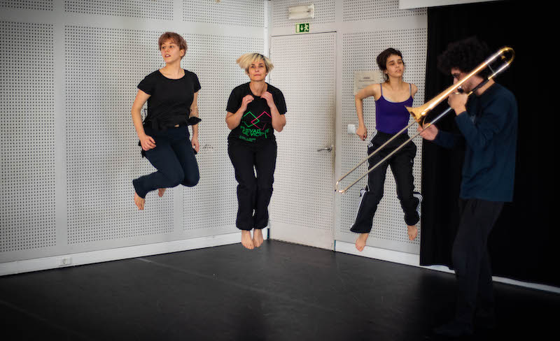 Na imagem estão quatro pessoas numa sala de ensaio. De frente para nós, três mulheres captadas num momento de salto, em que o seu corpo se suspende do ar. À direita um homem de perfil para a esquerda, que toca um trombone de varas.