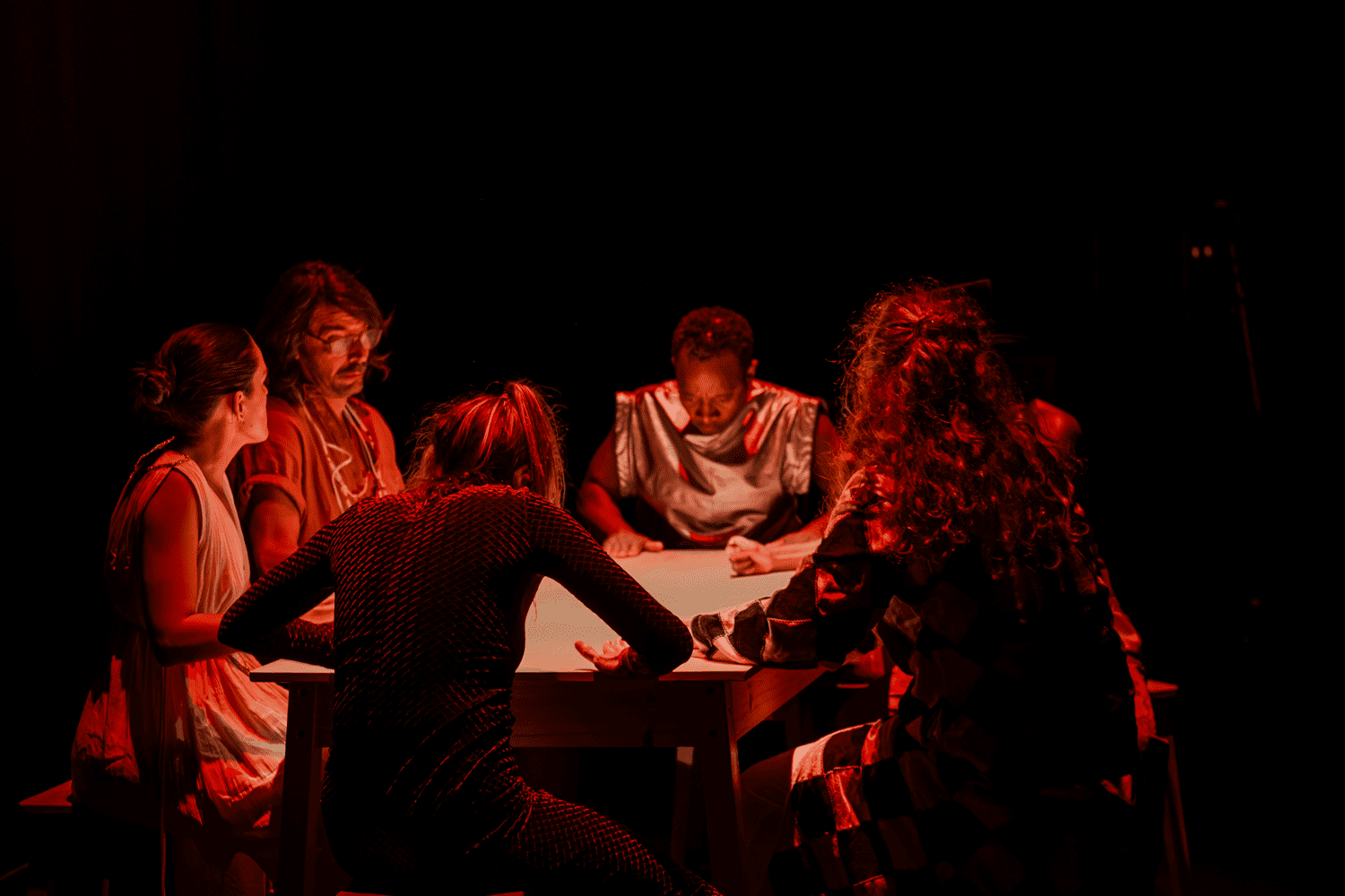 Seis intérpretes, reunidos à volta de uma mesa, participam numa cena iluminada por uma intensa luz vermelha.
